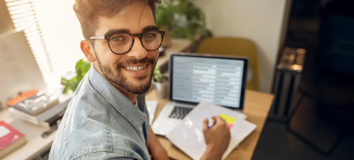 Smiling man with glasses at a desk, holding documents, looks at a laptop displaying code.