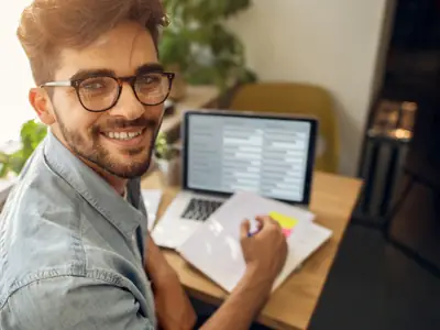 Smiling man with glasses at a desk, holding documents, looks at a laptop displaying code.