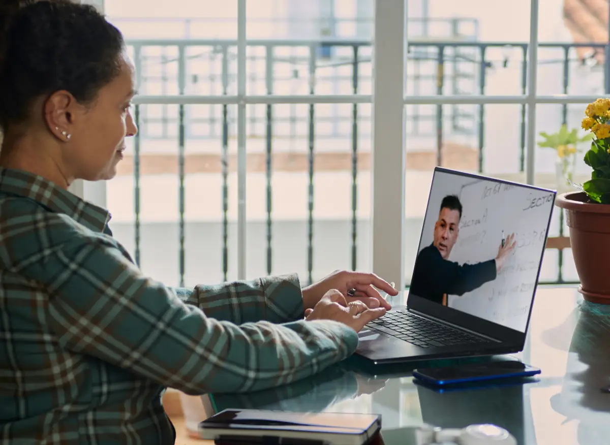A woman in a plaid shirt attentively watches an online lesson on her laptop.