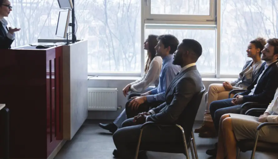 A group of students sit attentively in a classroom, facing a speaker at a podium. The room is well-lit by a large window, creating a professional and focused atmosphere.