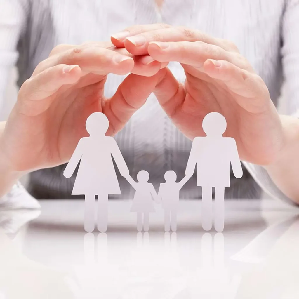 Hands hovering protectively over paper cutouts of a family, including two adults and two children, conveying care and security. The tone is warm and protective.
